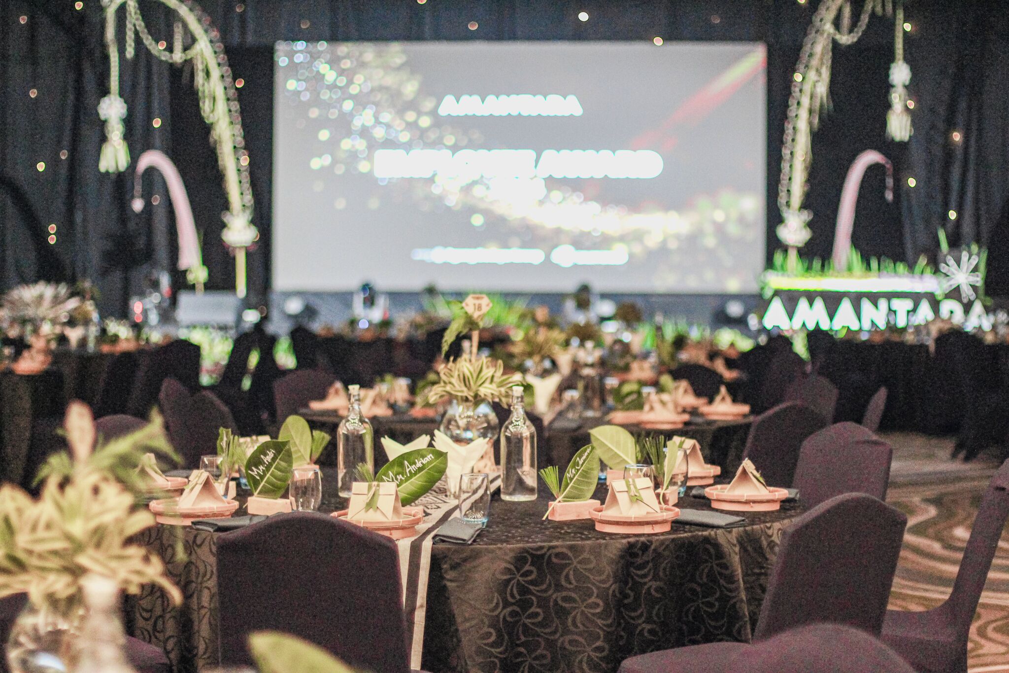 A banquet hall with tables set for a formal event, decorated with plants and glassware.