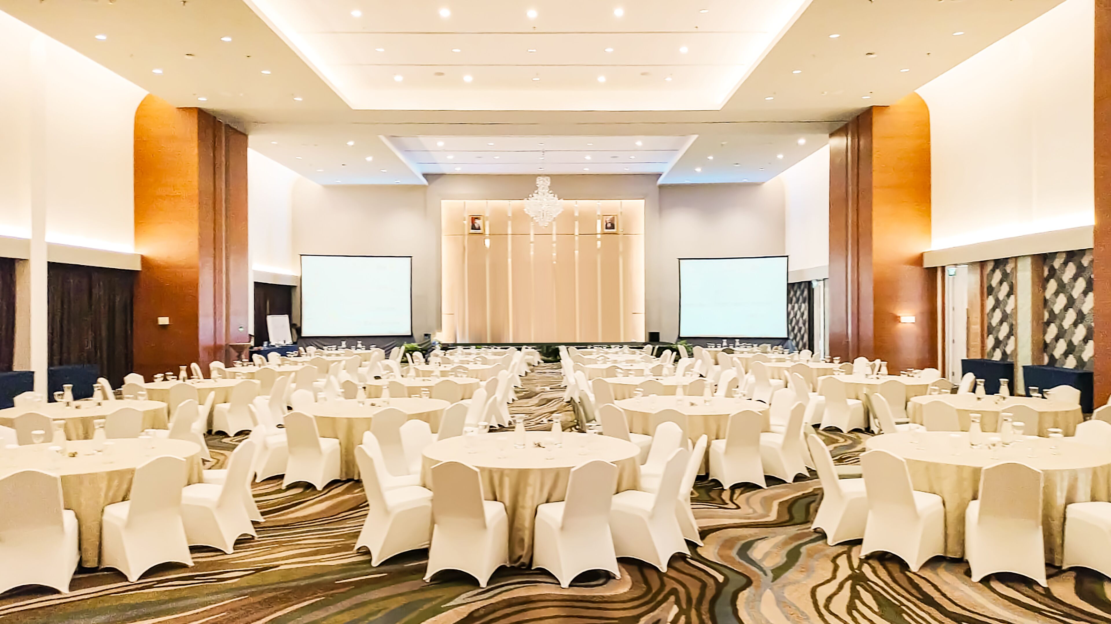 A large conference room with multiple round tables and white chairs, equipped with screens and a stage at the front.