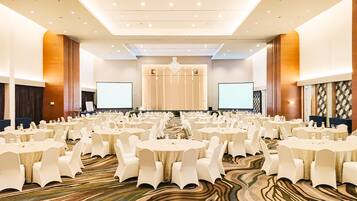 A large conference room with multiple round tables and white chairs, equipped with screens and a stage at the front.