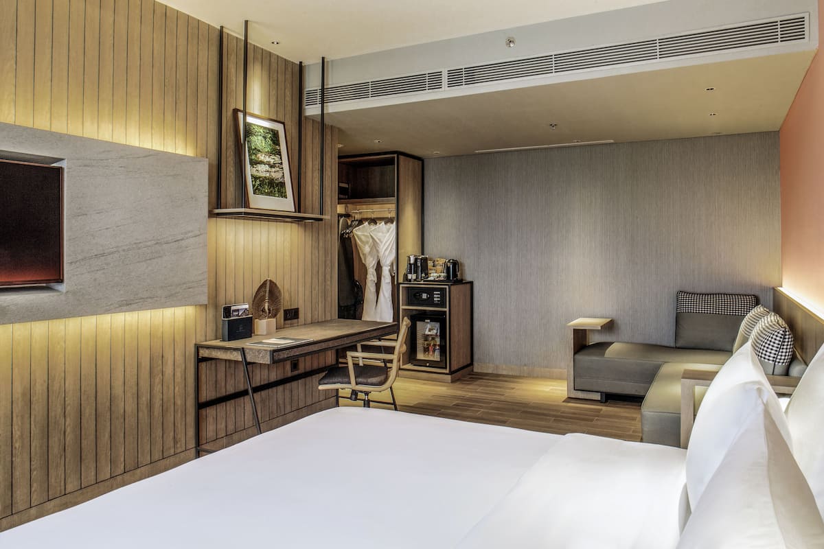 A modern hotel room with a bed, a desk, a chair, a sofa, and a wardrobe.