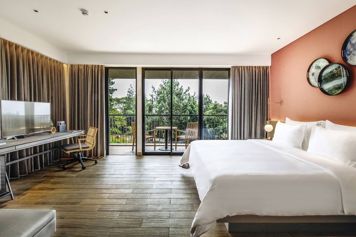 A hotel room with a large bed, a desk with a TV, and a balcony with a view of trees.