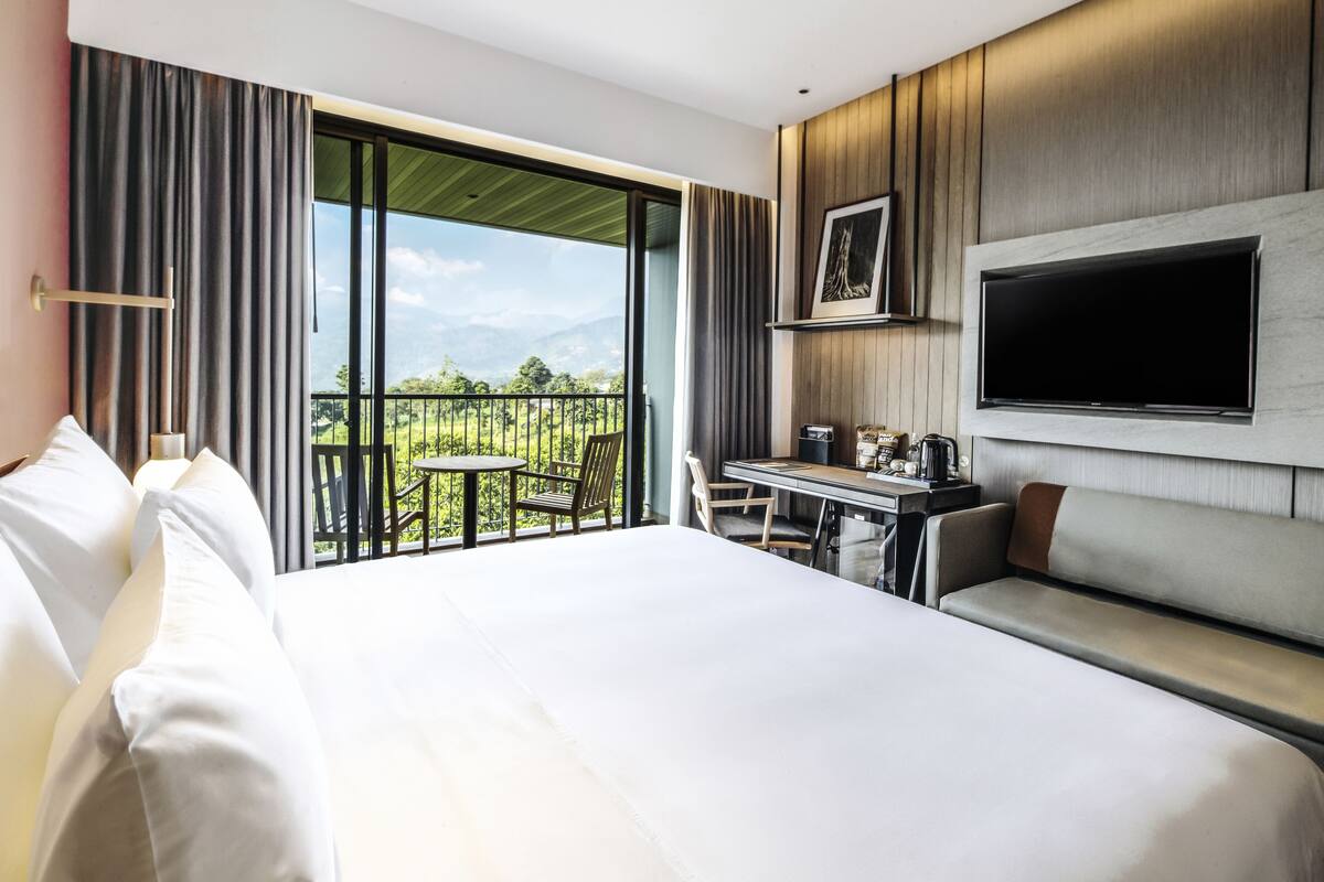 A hotel room with a large bed, a desk with a chair, a TV mounted on the wall, and a balcony with a view of the mountains.