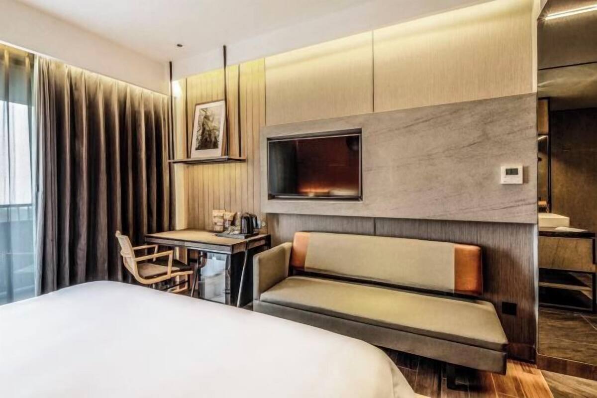 A hotel room with a bed, a desk with a chair, a sofa, a TV mounted on the wall, and a window with curtains.