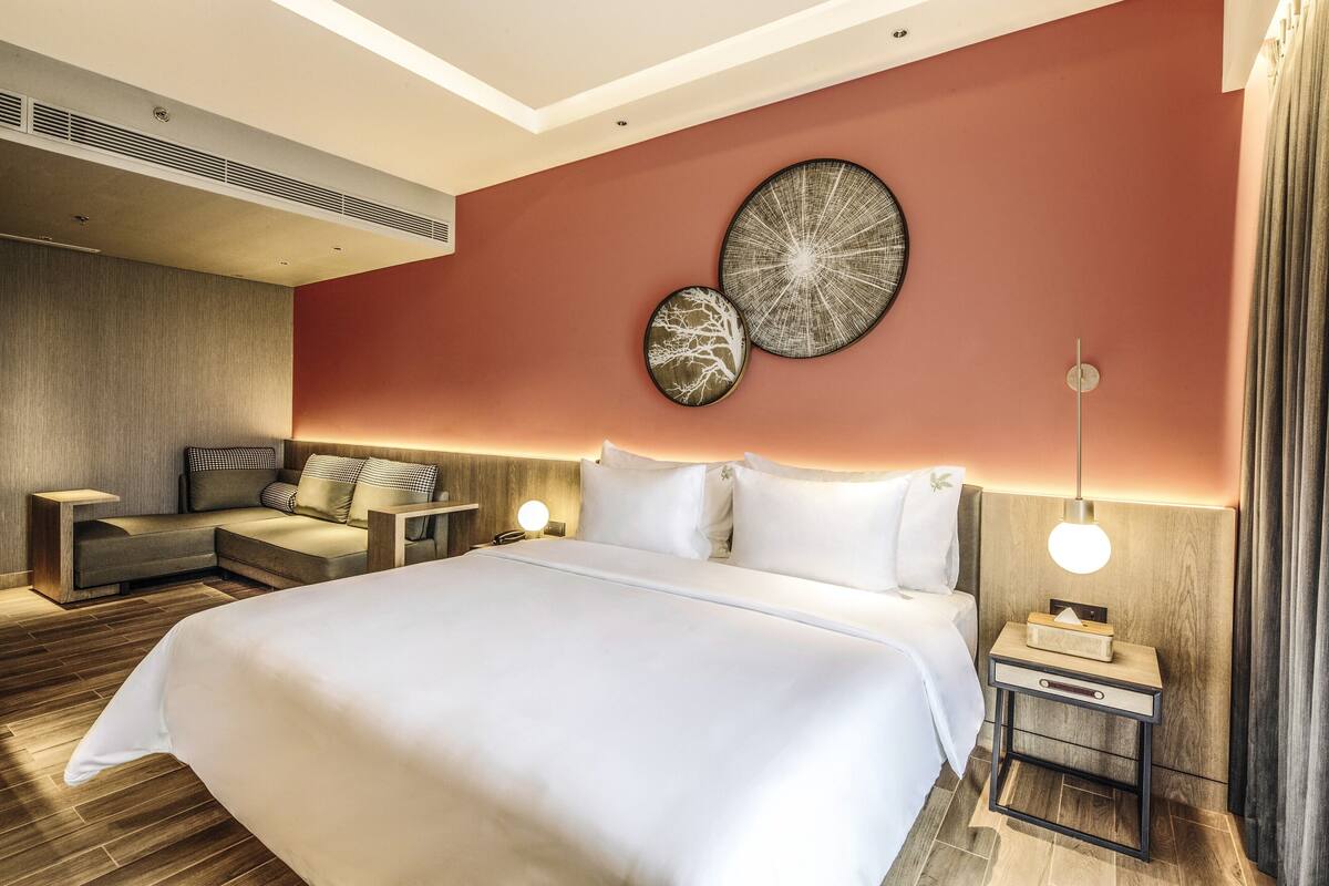 A modern hotel room with a large bed, a sofa, a nightstand with a lamp, and two wall art pieces.