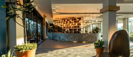 Un lobby moderno con una mesa de recepción, sillas de madera, plantas en macetas y un piso con diseño geométrico.