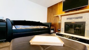 A modern living room with a black leather sofa, a flat-screen TV mounted on the wall, and a fireplace with a stone facade.