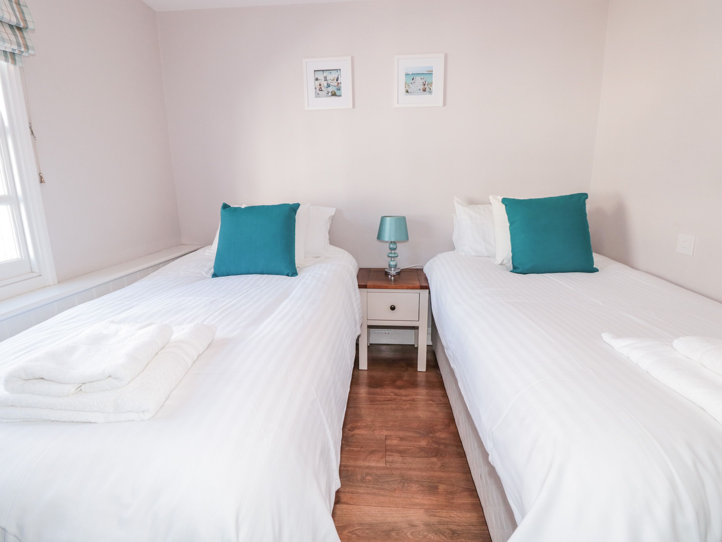 Hotellihuone, jossa on kaksi sänkyä, yöpöytä, lamppu ja kaksi kehystettyä kuvaa seinällä.