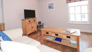 Un salon comprenant un canapé, une table basse en bois, un téléviseur posé sur un meuble, une fenêtre avec un rideau à carreaux et un tableau accroché au mur.