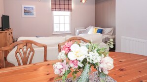 Une pièce chaleureuse avec une table à manger en bois, deux chaises en bois, un lit avec une couette blanche, une fenêtre avec des rideaux à carreaux et un téléviseur fixé au mur.