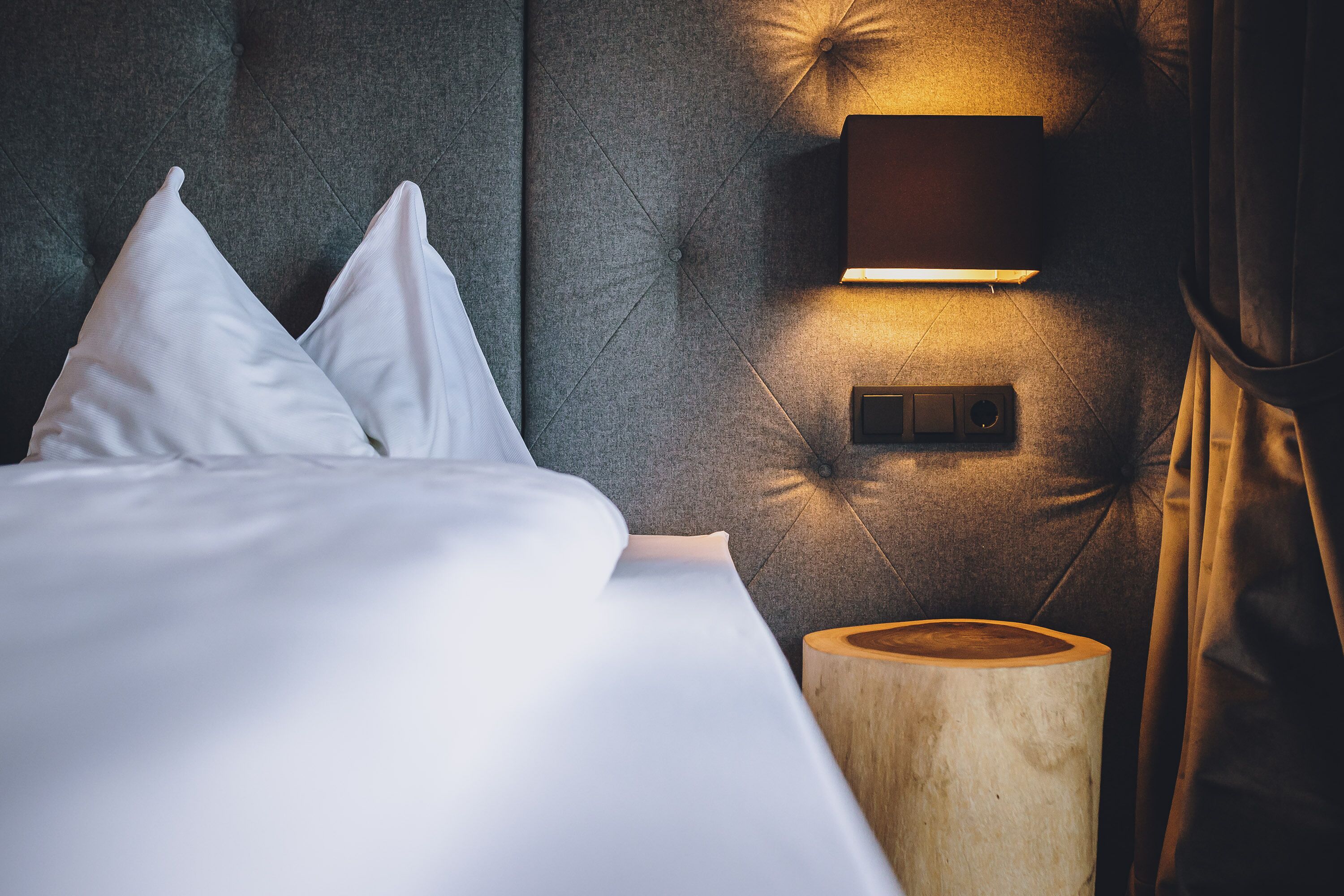Un lit bien fait, avec du linge de lit blanc et des oreillers, une tête de lit capitonnée, un luminaire fixé au mur et une table de chevet en bois.