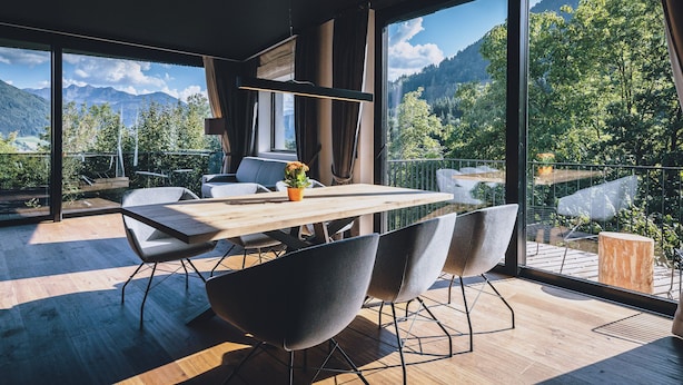 Un espace repas moderne avec une table et des chaises en bois, de grandes fenêtres offrant une vue sur les montagnes et un balcon doté d’une terrasse en bois.