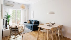 A modern living room with a blue sofa, wooden dining table, and chairs. There is a large window with curtains, a hanging light fixture, and a potted plant. - Absolute 2BR Apartment (Athens)