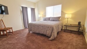 Een slaapkamer met een bed, een stoel, een aan de muur bevestigde televisie en een raam met gordijnen.
