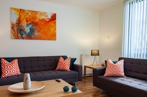Una sala de estar moderna con dos sofás oscuros, cojines decorativos estampados, una mesa de centro de madera y un cuadro abstracto grande en la pared.