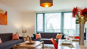 A modern living room with a sofa, coffee table, and dining area. There is a large window with blinds, a vase with flowers, and a sign for 'BYEVO Serviced Apartments'. - ByEvo 5 Brabloch GLA Apartment  (Paisley)