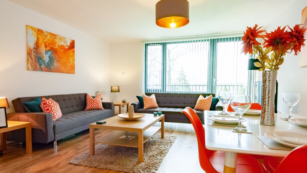 A modern living room with a grey sofa, red and orange cushions, a wooden coffee table, and a dining area with a white table and red chairs. - ByEvo 5 Brabloch GLA Apartment  (Paisley)