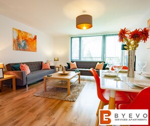 Ein modernes Wohnzimmer mit grauem Sofa, roten und orangefarbenen Kissen, einem Holztisch und einem Essbereich mit weißem Tisch und roten Stühlen.