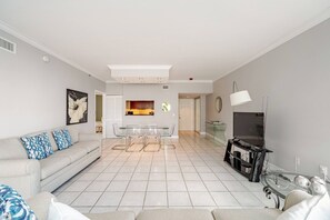 A modern living room with a sofa, a dining table with chairs, a TV, and a kitchen area in the background.