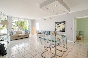 A spacious living room with a glass coffee table, two sofas with patterned cushions, a large abstract painting on the wall, and a view of palm trees through the large windows.