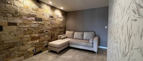 Un moderno salón con una pared de acento de piedra, un sofá seccional color beige y suelo de baldosas.