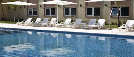 Un espace aménagé au bord de la piscine, avec des chaises longues et des parasols, une piscine à eau claire et un bâtiment doté de nombreuses fenêtres.
