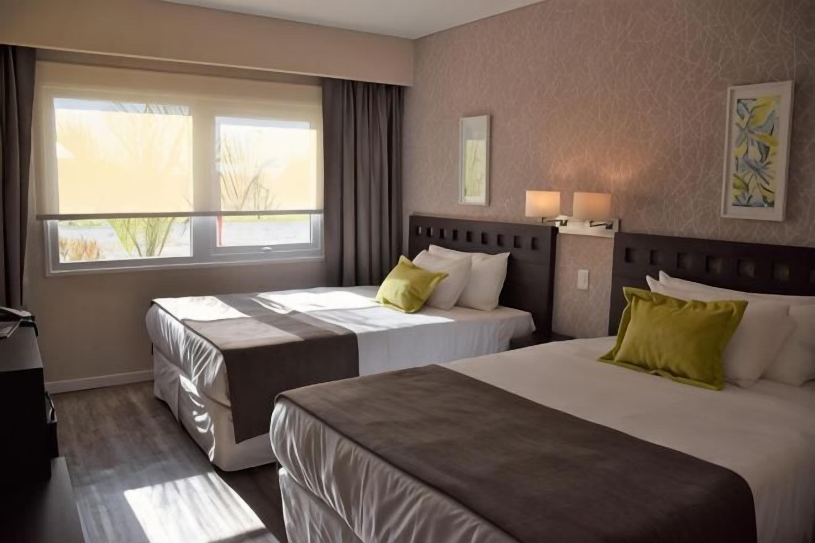 A hotel room with two beds, a window with curtains, a nightstand with lamps, and a painting on the wall.