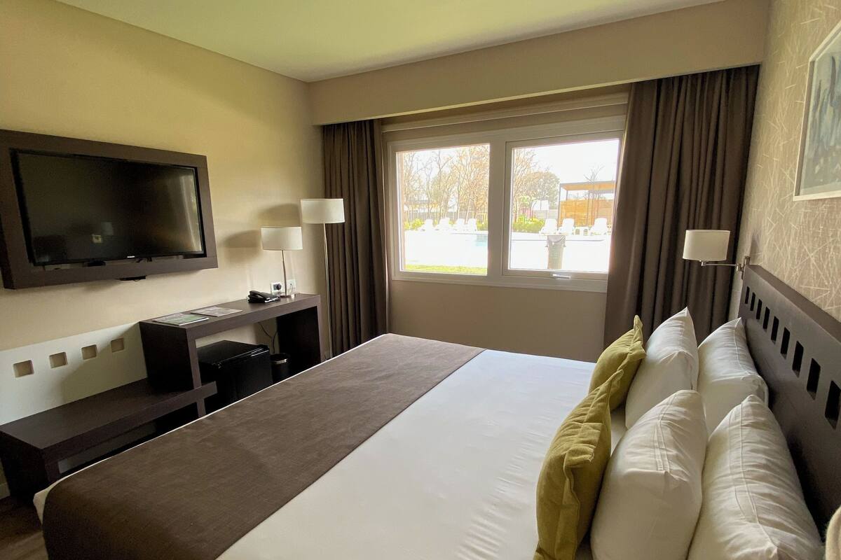 A hotel room with a large bed, a desk, a flat-screen TV mounted on the wall, and a window with curtains.
