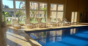 An indoor swimming pool with clear glass walls, multiple lounge chairs, and a view of a landscaped area with buildings in the background.