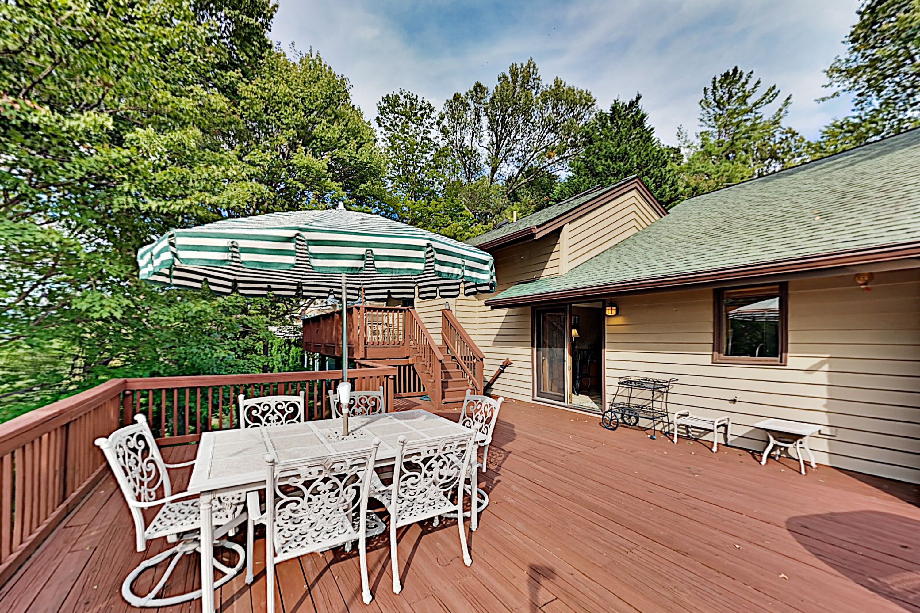 A wooden deck with a table and chairs, a green and white striped umbrella, and a staircase leading to a building.
