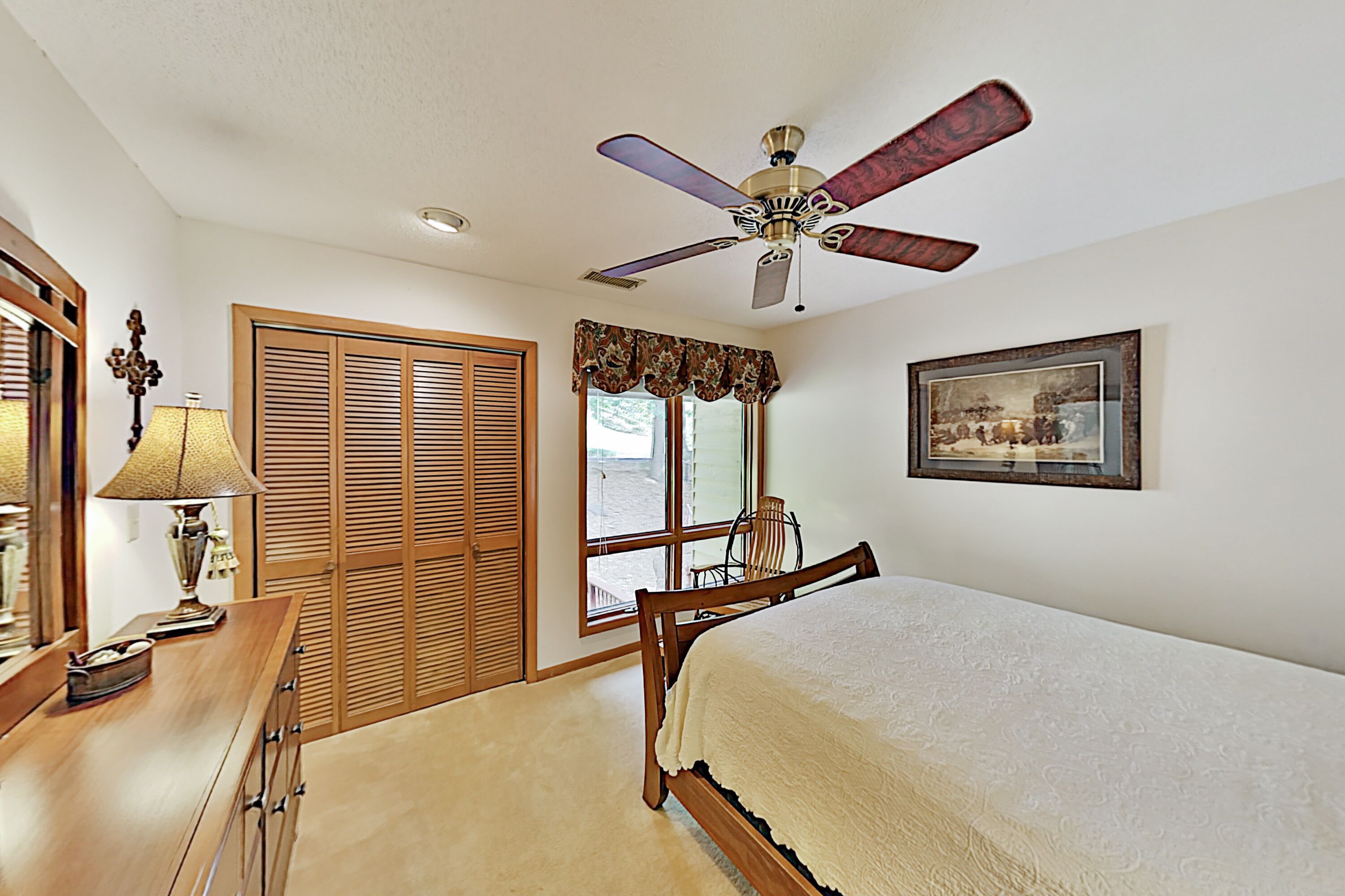 A bedroom with a bed, wooden furniture, a ceiling fan, a window with a view, and a framed picture on the wall.