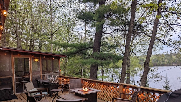 A wooden deck with outdoor furniture, a fire pit, and a view of a lake surrounded by trees.