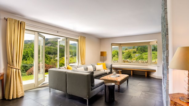Ein modernes Wohnzimmer mit einem grauen Sofa, einem Holztisch und einer Bank am Fenster. Große Fenster bieten Blick auf Grünflächen und Hügel.