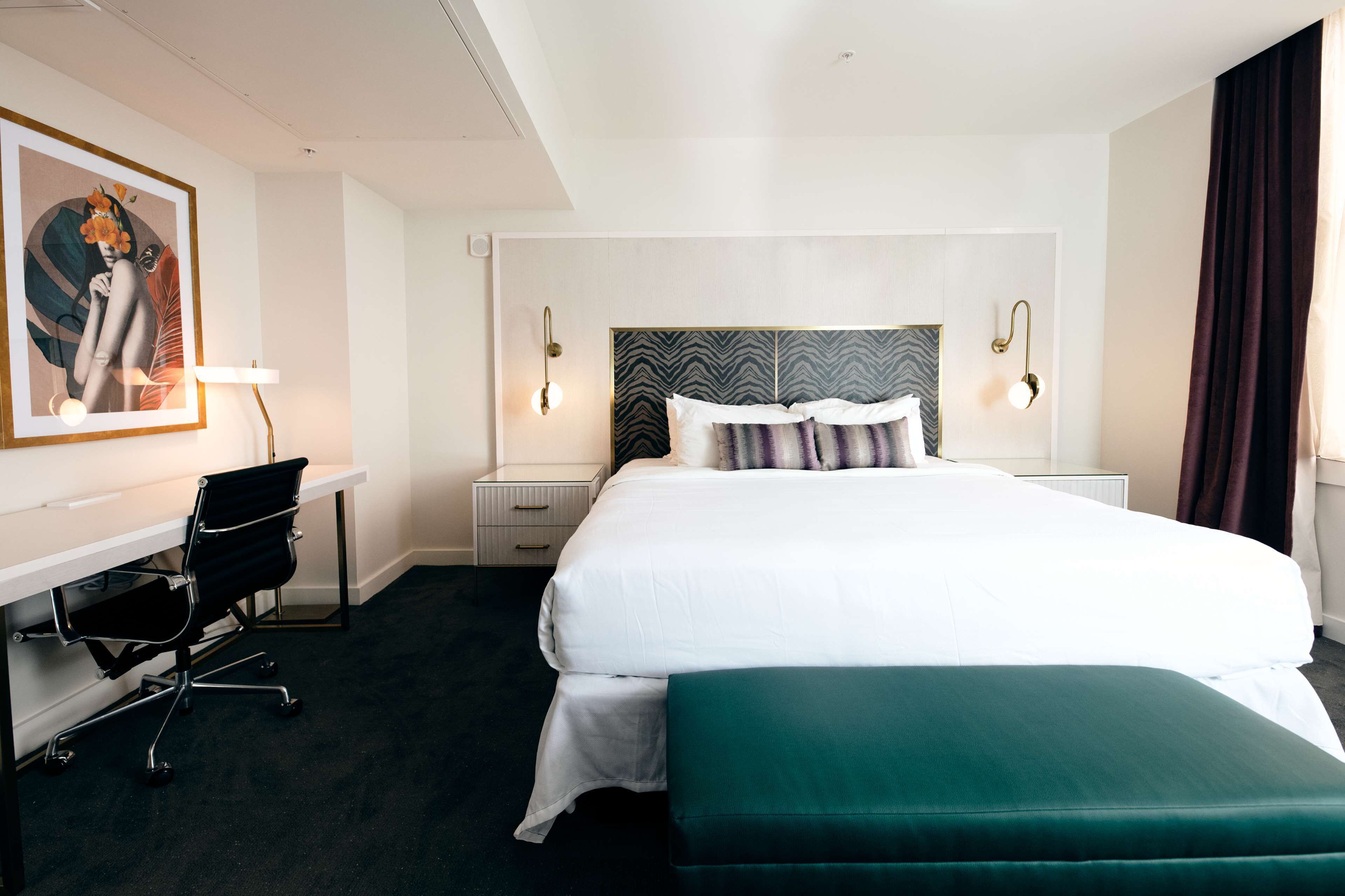 A hotel room with a large bed, a desk with a chair, a painting on the wall, and two bedside lamps.