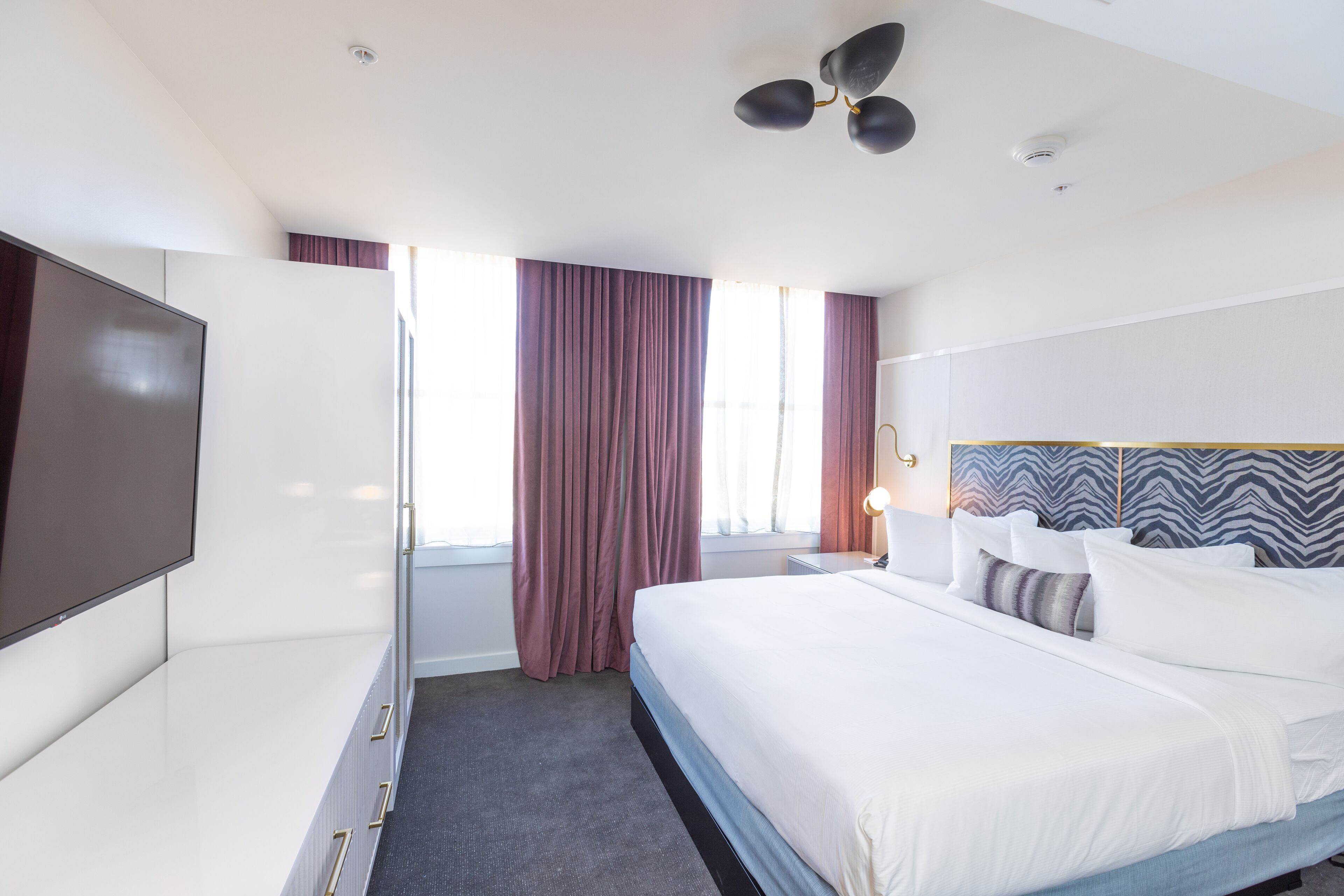 A hotel room with a bed, a TV, a ceiling fan, and a window with curtains.