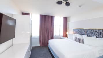 A hotel room with a bed, a TV, a ceiling fan, and a window with curtains.