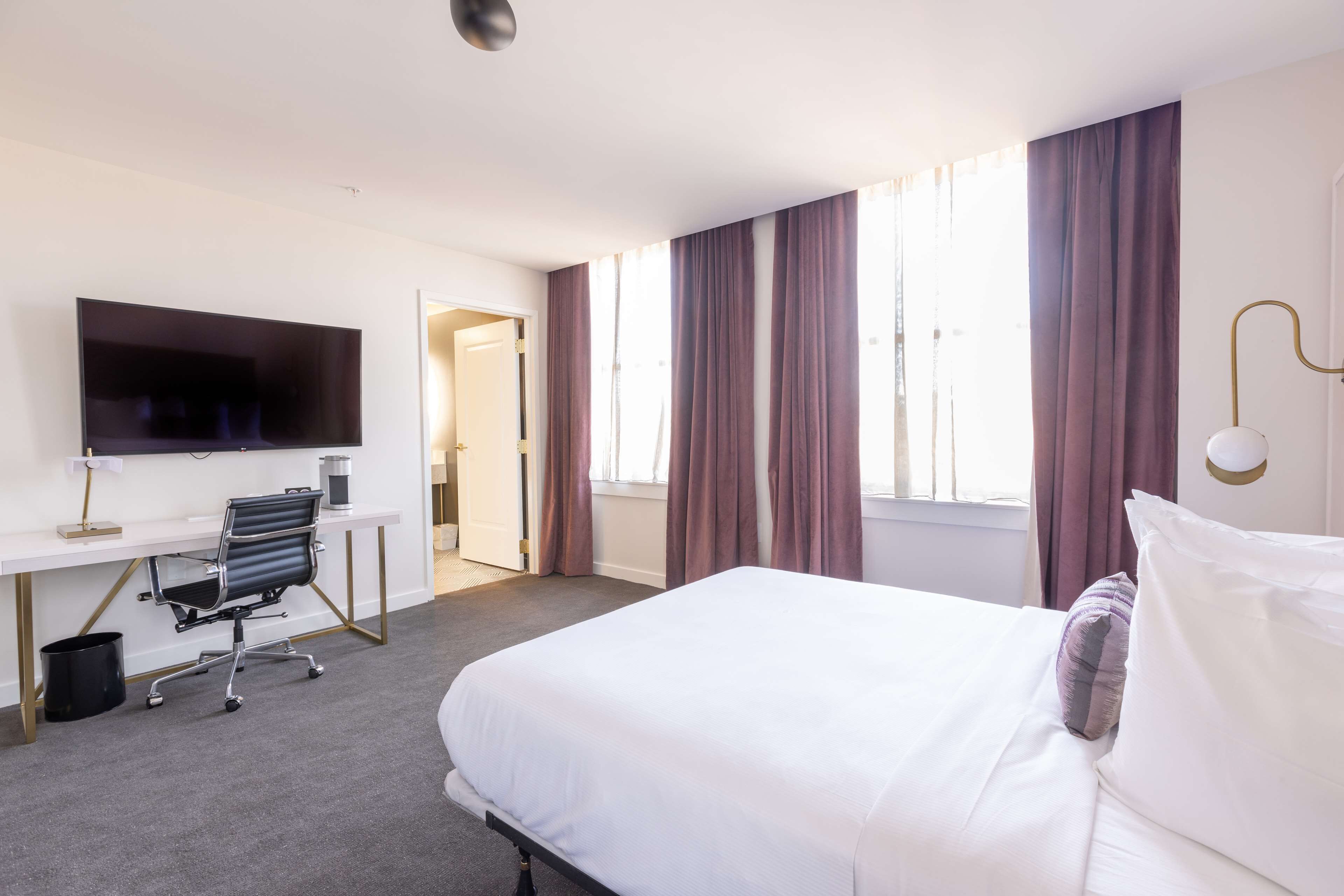 A hotel room with a bed, a desk with a computer, a TV, and a window with curtains.