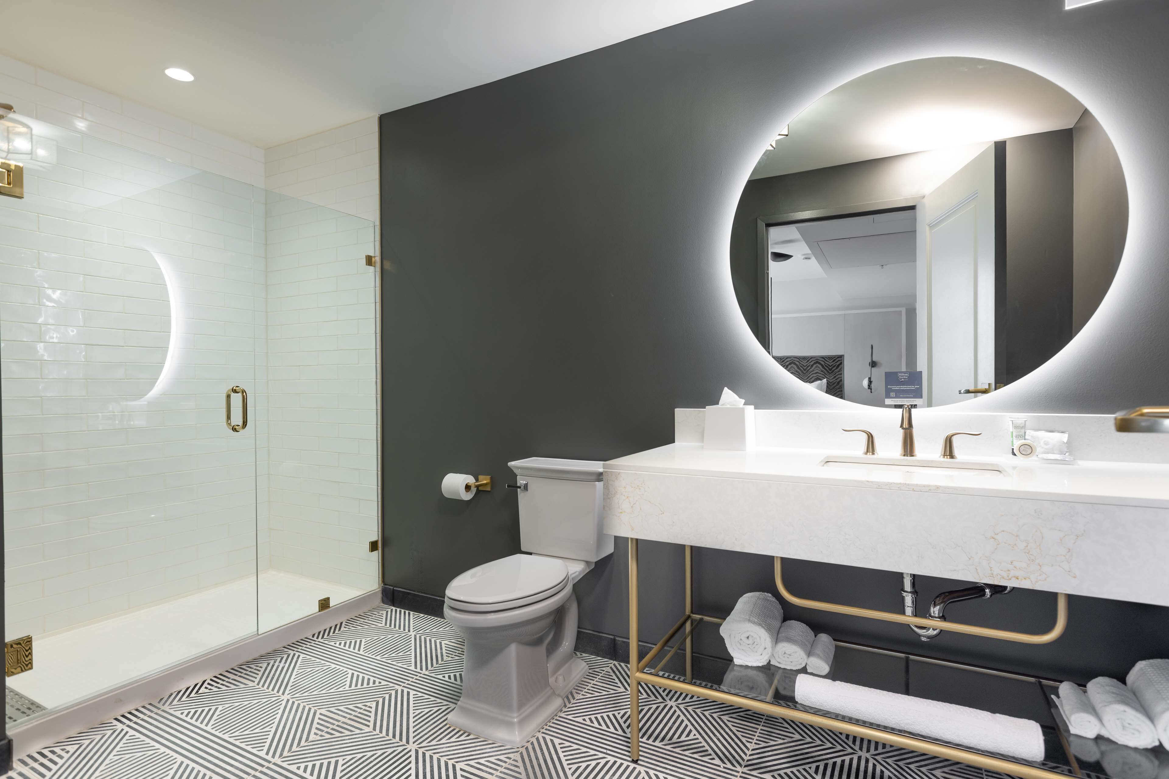 Un baño moderno con cabina de ducha de vidrio, inodoro, tocador con un espejo grande y suelo estampado.