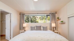 Ein Schlafzimmer mit einem großen Bett, zwei Nachttischen mit Lampen, einem Fenster mit Vorhängen und Blick ins Grüne.