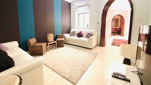 Una sala de estar con un sofá blanco, un sillón marrón y una mesita con una lámpara. Hay un televisor sobre un mueble bajo y una puerta que da a otra habitación.