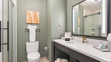 A modern bathroom with a white toilet, a glass shower enclosure, a white sink with a gold faucet, and a large mirror.