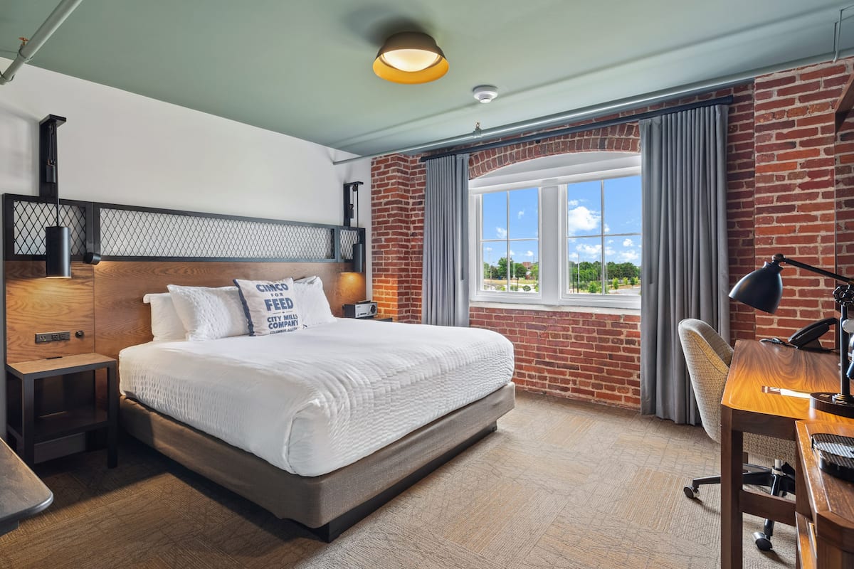 A hotel room with a large bed, a desk with a chair, a window with a view of greenery, and a brick wall.