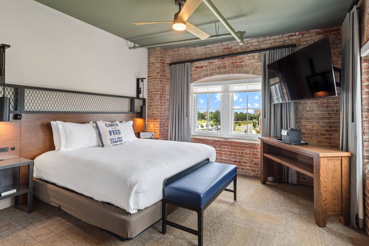 A hotel room with a bed, a bench, a desk, a TV, and a window with a view.