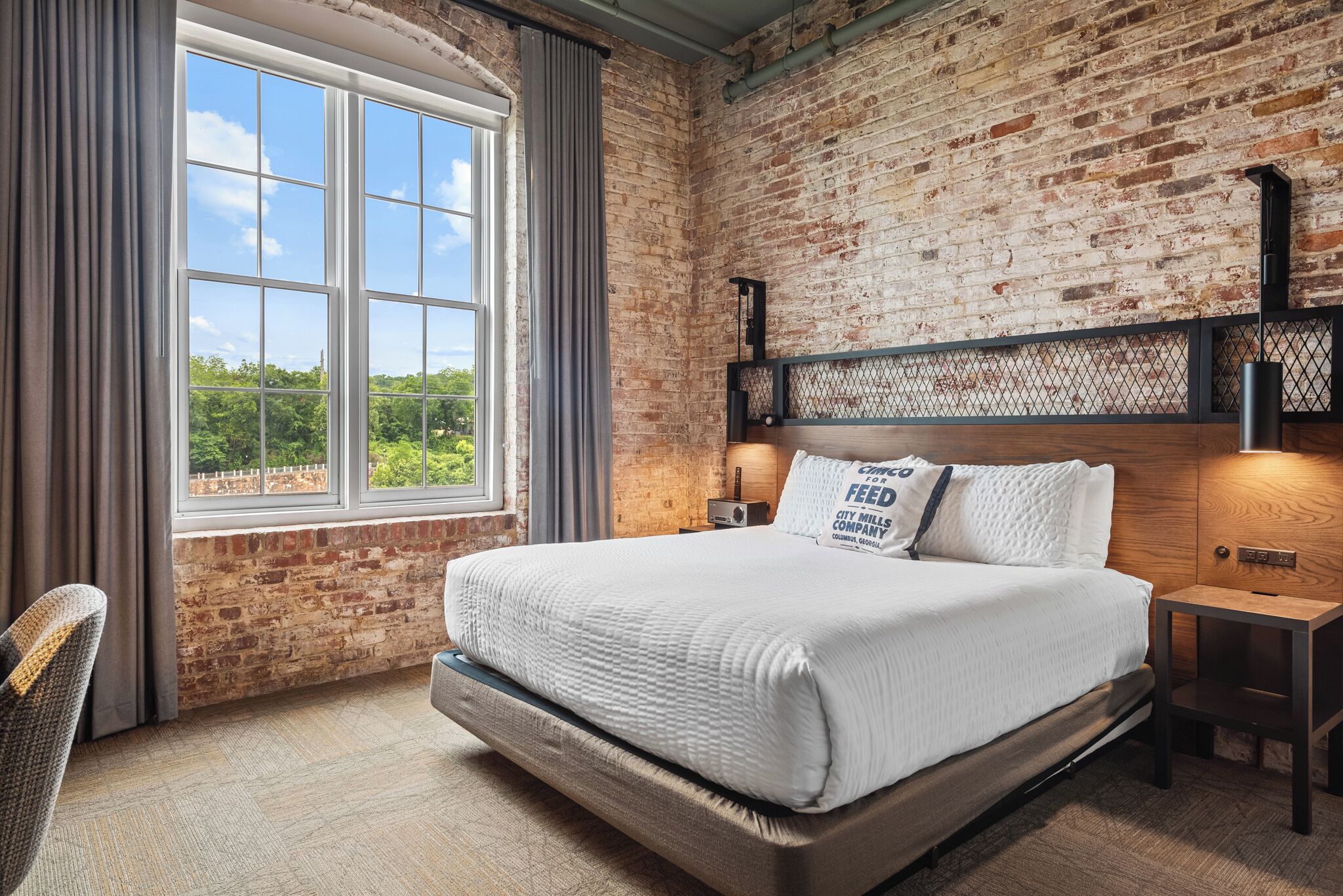 A bedroom with a brick wall, a large window, a bed with white bedding, and a nightstand with a lamp.