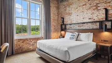 A bedroom with a brick wall, a large window, a bed with white bedding, and a nightstand with a lamp.