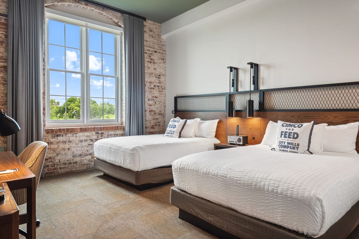 A hotel room with two beds, a desk, a chair, a window with curtains, and a brick wall.