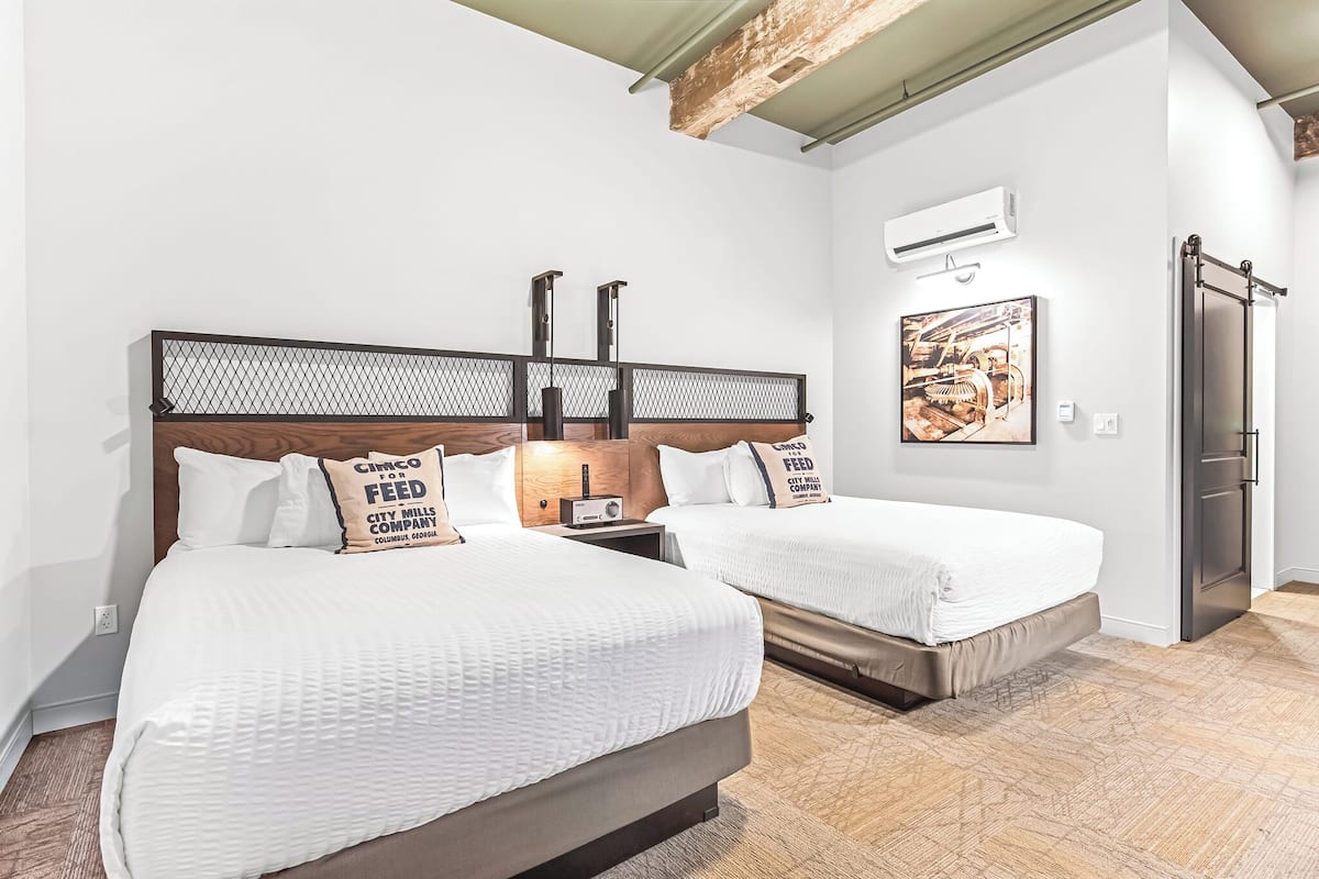 A hotel room with two beds, a wooden headboard, a wall-mounted air conditioner, and a framed picture.