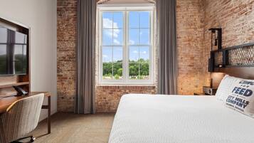 A hotel room with a brick wall, a large window with curtains, a bed with a white comforter, a desk with a chair, and a television on the wall.