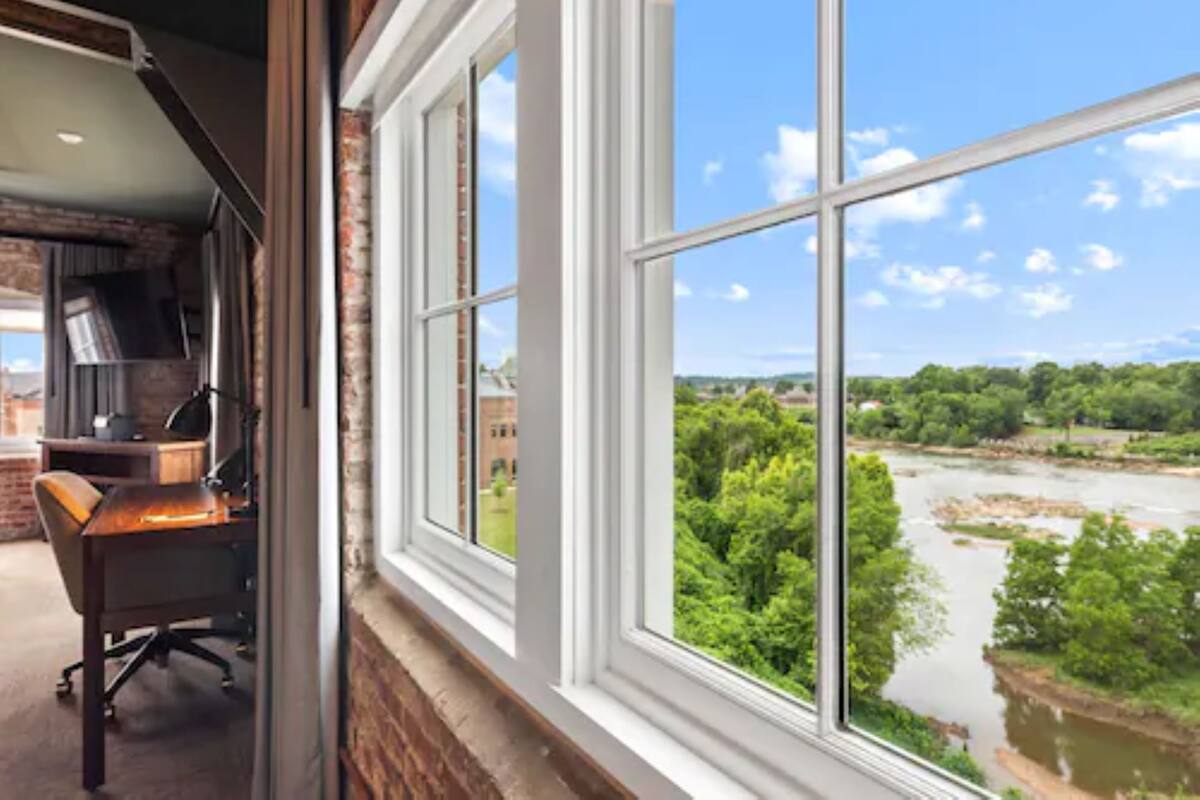 A room with a brick wall, a desk, and a large window overlooking a river and greenery.