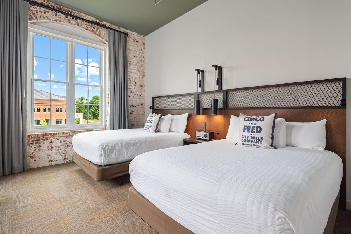 A hotel room with two beds, a brick wall, a large window, and a view of a building outside.