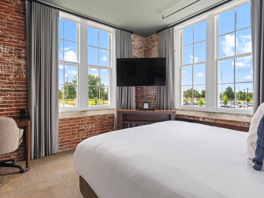 Quarto de hotel com parede de tijolos, janelas amplas, televisão de tela plana, cama, escrivaninha e uma cadeira.